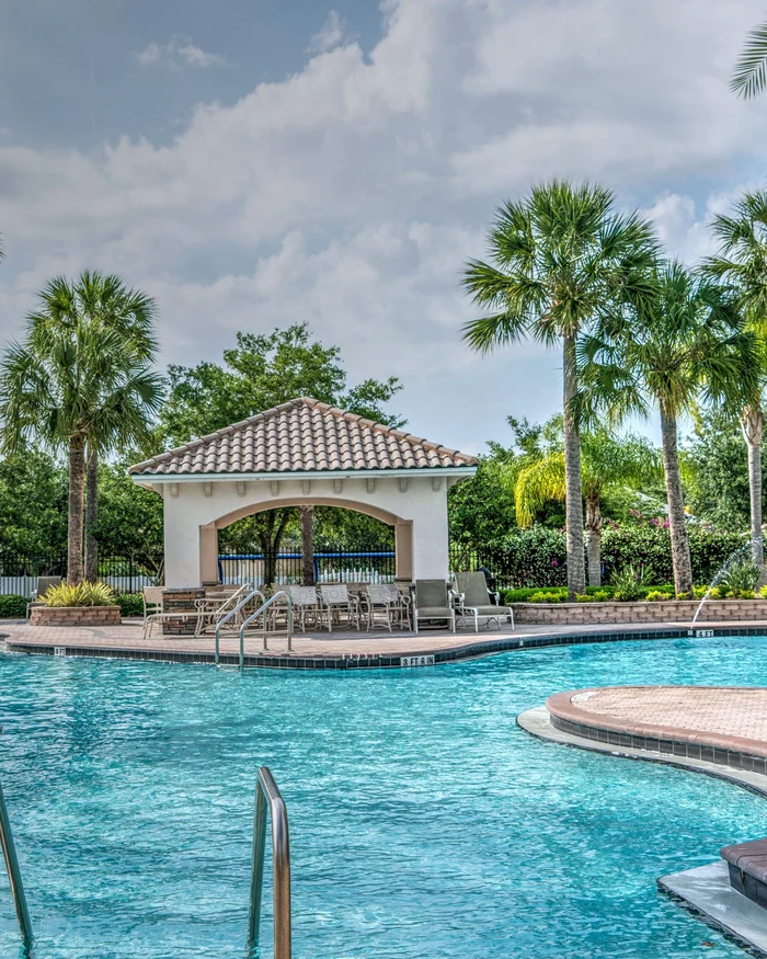 A large community pool with a pool deck, lounge chairs and a gazebo under palm trees